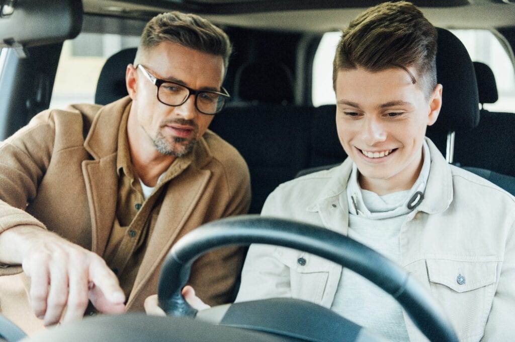 handsome father pointing on something while teaching smiling teen son driving car